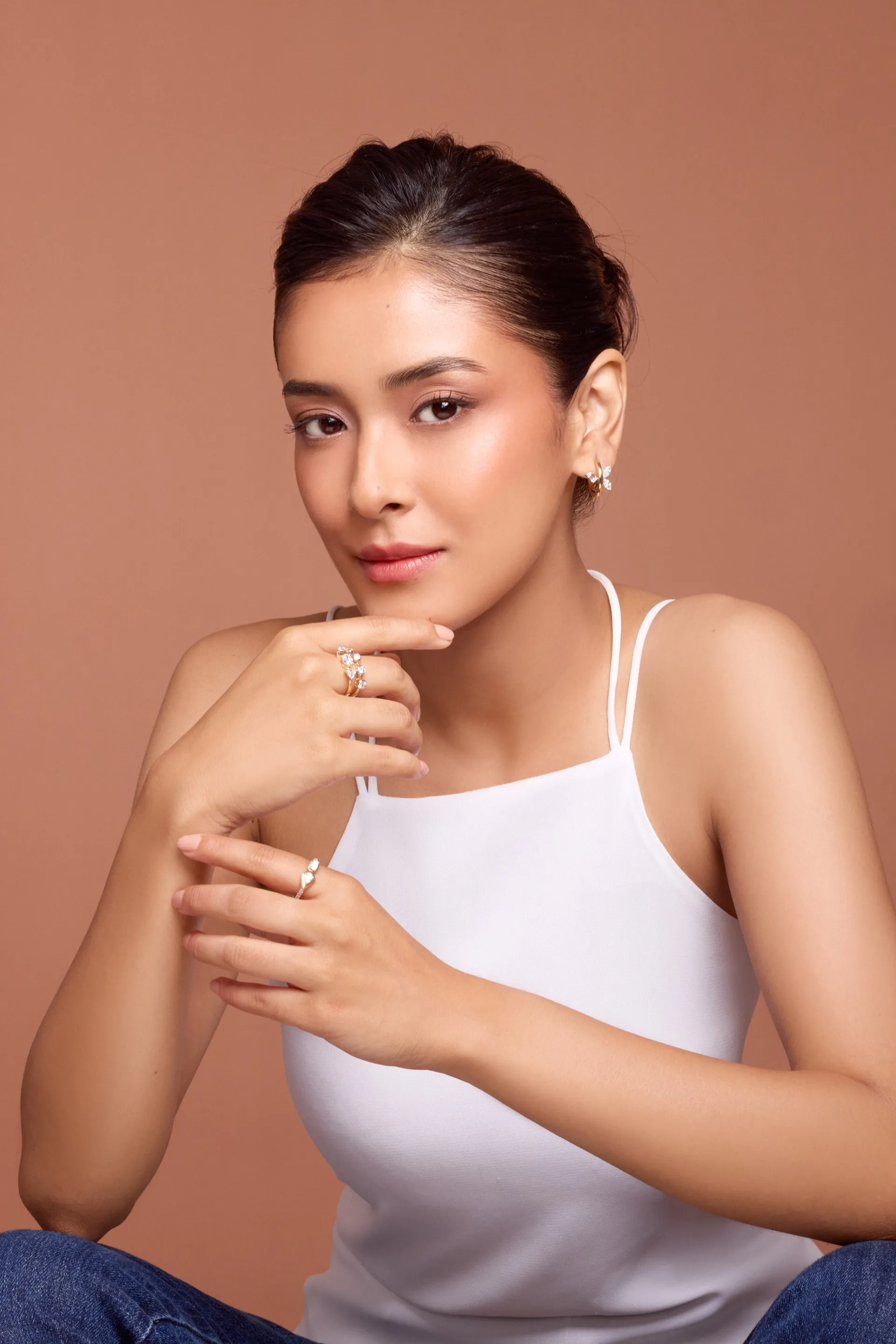 Portrait of a woman in a white top wearing Tuisa diamond rings and stud earrings, seated against a terracotta backdrop