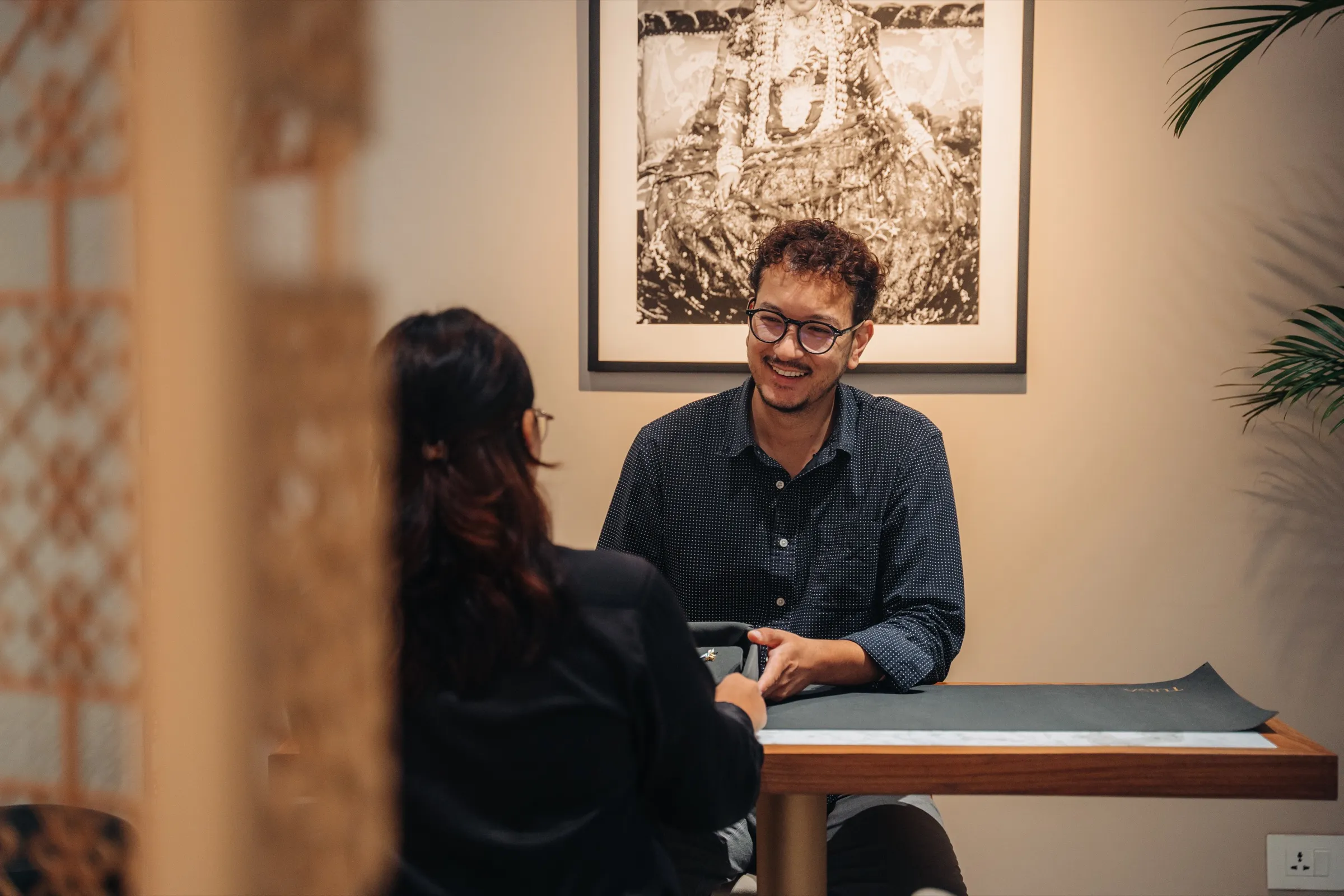 A Tuisa consultation: two people in conversation at the table beneath an archival portrait, seen past the brass jaali screen