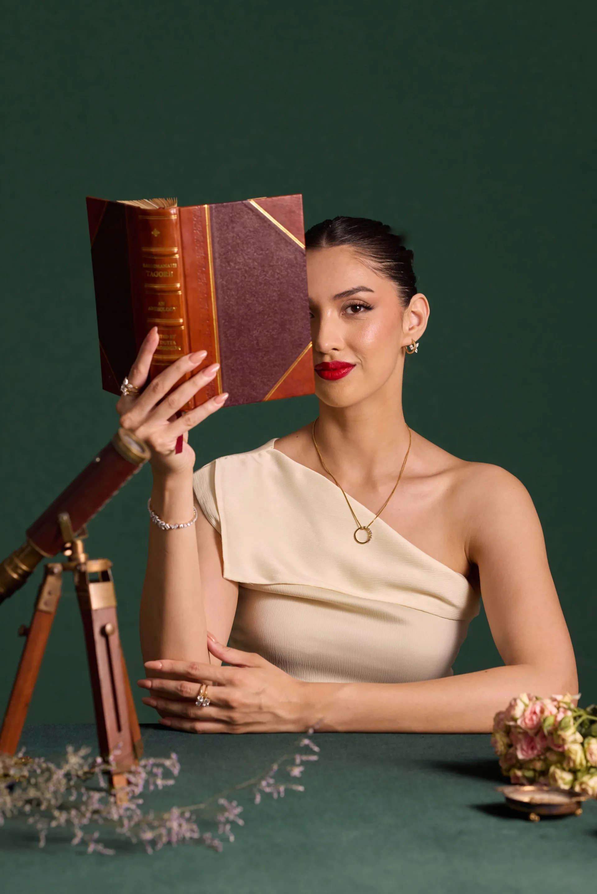 Editorial portrait of a woman holding an antique book over one eye, wearing Tuisa jewellery against a deep green backdrop